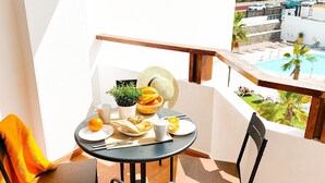Een balkon met een tafel gedekt voor ontbijt, inclusief sinaasappelschijfjes, een croissant en een fruitschaal. Op tafel staat een stoel en een zonnehoed.