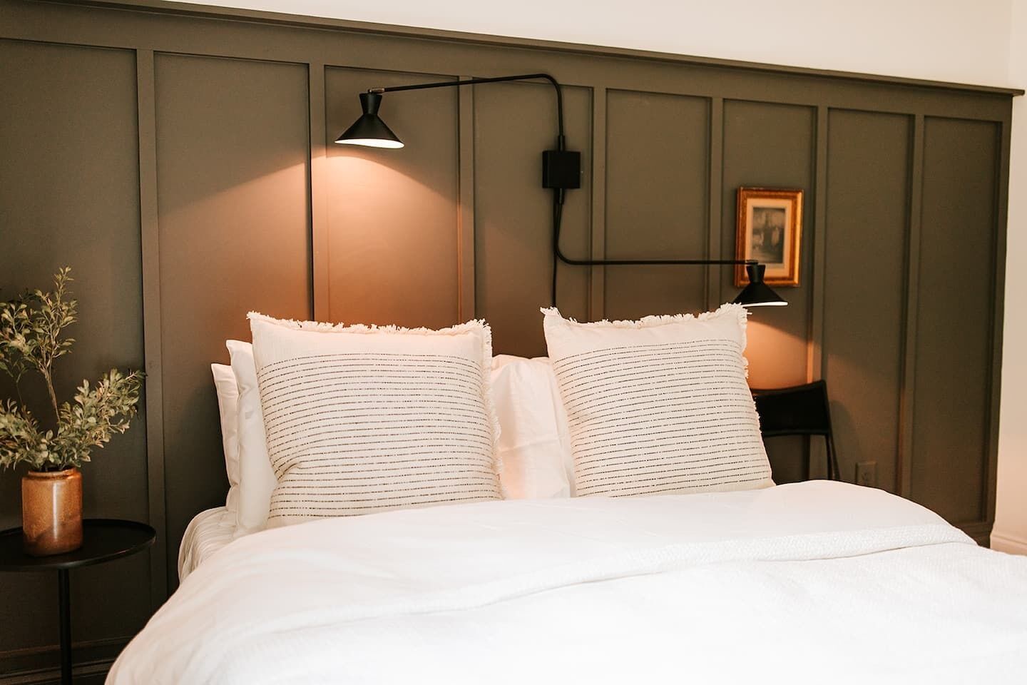 Een netjes opgemaakt bed met twee kussens, een bedlampje, een tafeltje met een plantje en een lijstje aan de muur.