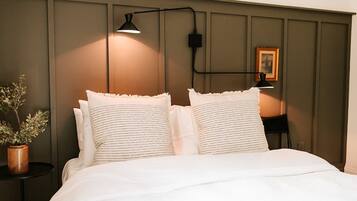 Een netjes opgemaakt bed met twee kussens, een bedlampje, een tafeltje met een plantje en een lijstje aan de muur.