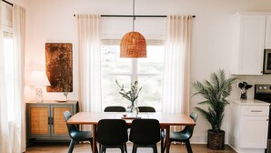 Un espace repas avec une table en bois, des chaises noires, un luminaire suspendu en fibres tressées et une vue sur l’extérieur grâce à de grandes fenêtres.