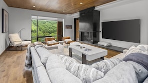 Relax in a spacious living room with a sofa, chairs, and a flat-screen TV, offering a view of the lush green landscape.