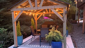 A wooden gazebo with a table and chairs, surrounded by potted plants and a patterned rug. - Modern studio with hot tub and patio access  (Poulsbo)