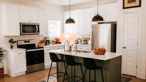 Una cocina moderna con armarios blancos, electrodomésticos de acero inoxidable, una isla central con taburetes y luminarias colgantes, con vista al área de comedor.