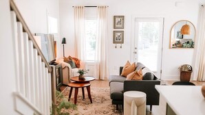 Una sala de estar moderna con un sofá, una mesa de centro y una pequeña alfombra. Se ve una escalera a la izquierda y una puerta con cortinas a la derecha.