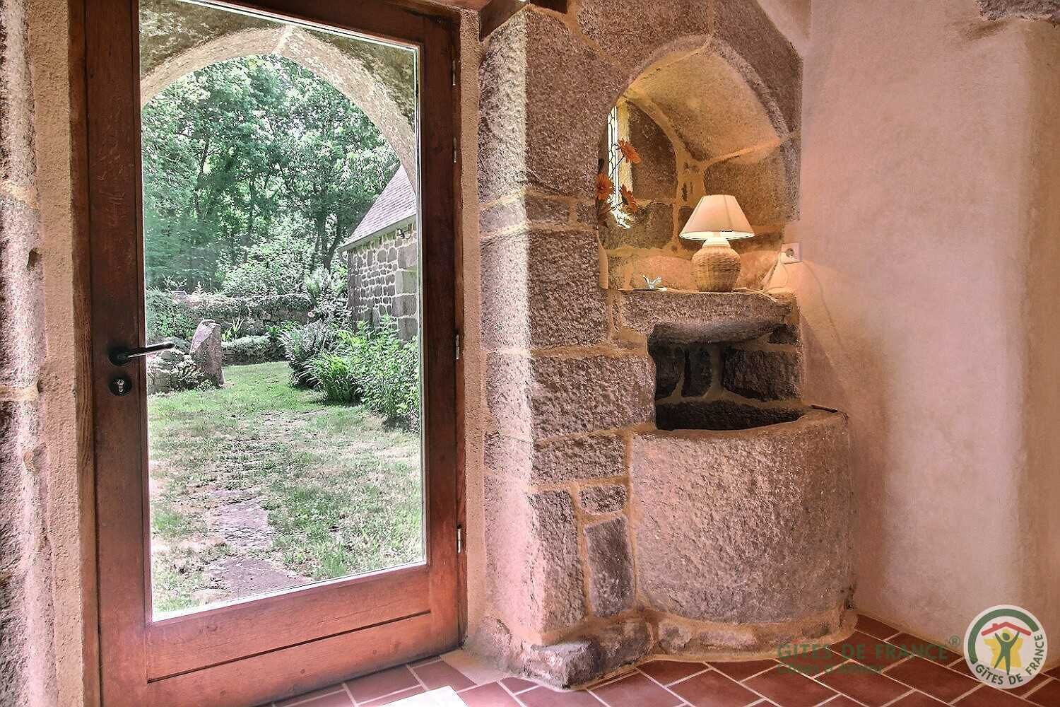 A stone-built room with a door leading to a garden, a stone fireplace with a lamp, and a tiled floor.