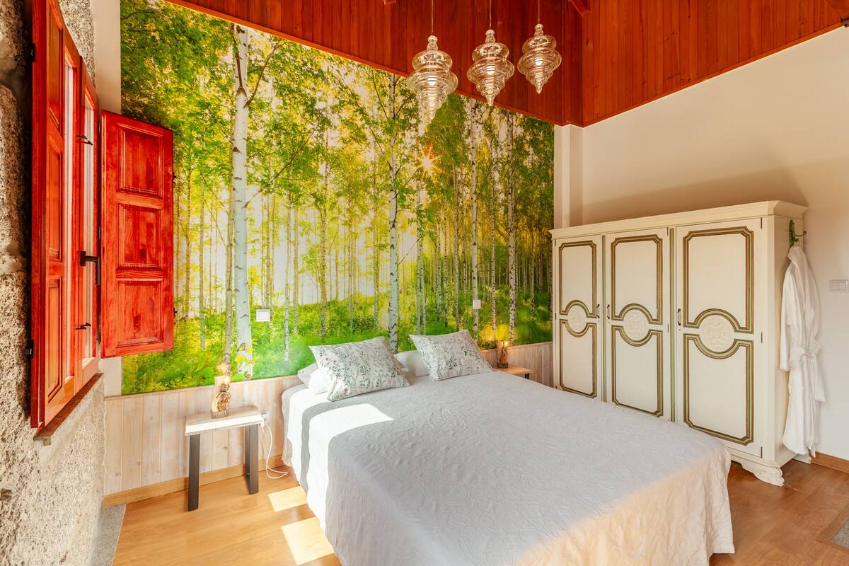 A bedroom with a large bed, a wardrobe, a small table, and a wall mural of trees.