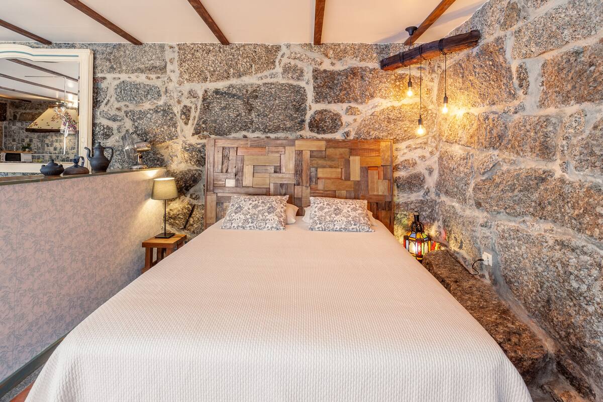 A bedroom with a stone wall, a wooden headboard, a bed with white bedding, and a nightstand with a lamp.
