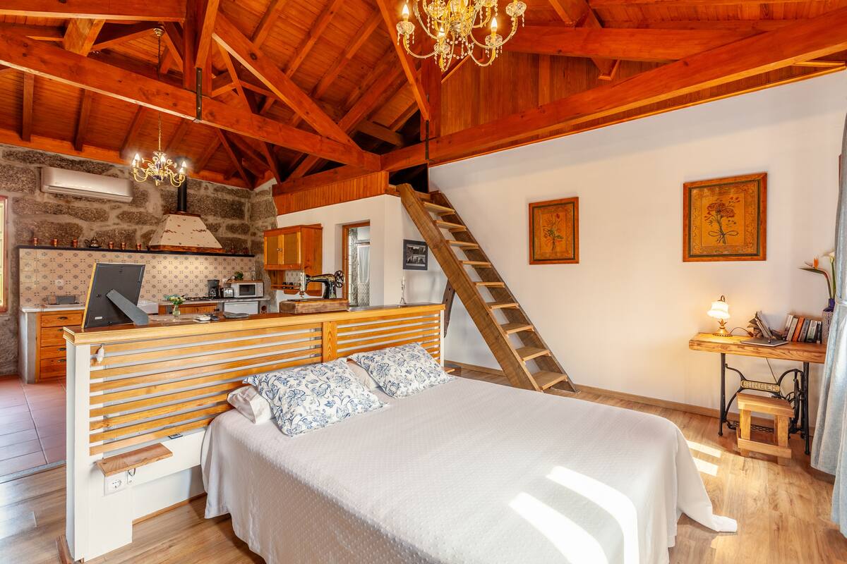 A bedroom with a wooden bed, a desk, a chair, a ladder, and a kitchenette in the background.