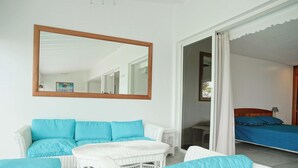 Una habitació amb un sofà i butaques de vimet blanc, un llit amb roba de llit blava i un mirall gran a la paret.