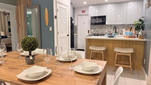 Un comedor con una mesa de madera dispuesta para cuatro, una pequeña planta en maceta y vistas a la cocina, con armarios blancos y salpicadero de azulejos.