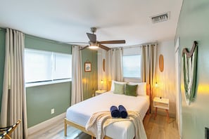 Habitación de hotel con cama, ventilador de techo, mesita de noche, lámpara y ventana con cortinas.