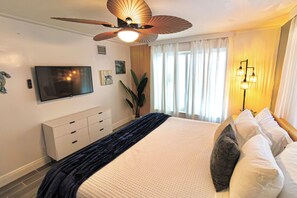 Ein Hotelzimmer mit einem Bett, einem Flachbildfernseher, einem Deckenventilator, einem Fenster mit Vorhängen und einem Nachttisch mit Lampe.