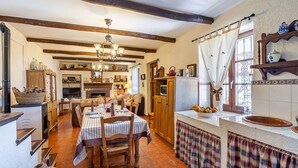 A cozy dining area with a long table set for four, wooden chairs, a chandelier, and a kitchen area with a tiled backsplash and wooden cabinets. - Cortijo La Alberquilla Íllora by Ruralidays (Granada)