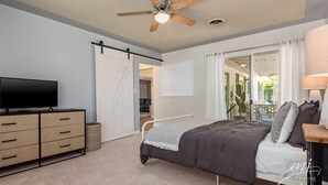A bedroom with a bed, a TV, a ceiling fan, a sliding door, and a view of the outdoors.