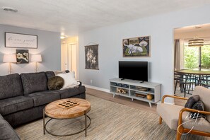 A modern living room with a grey sofa, a round wooden coffee table, a flat-screen TV, and a dining area with a table and chairs.