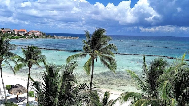 A beach with clear turquoise water, palm trees, and a distant shoreline with buildings. - PRIME Location, Gorgeous Sweeping Views, PH right on the Beach, Villas del Mar 1 (Puerto Aventuras)