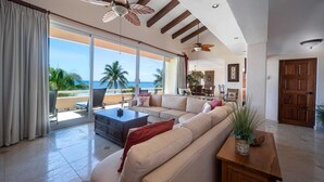 A spacious living room with a large sofa, a coffee table, and a view of the ocean through large sliding doors. - PRIME Location, Gorgeous Sweeping Views, PH right on the Beach, Villas del Mar 1 (Puerto Aventuras)