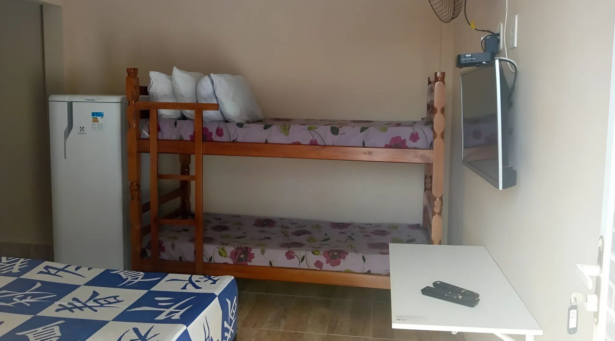Una cama individual con ropa de cama floral, un refrigerador pequeño, un escritorio con un control remoto y una cama con cubierta a cuadros.