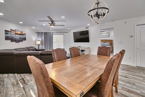 Un salon moderne avec une table à manger en bois, des fauteuils en cuir, un canapé en cuir marron, un ventilateur de plafond, un téléviseur à écran plat et un lustre.