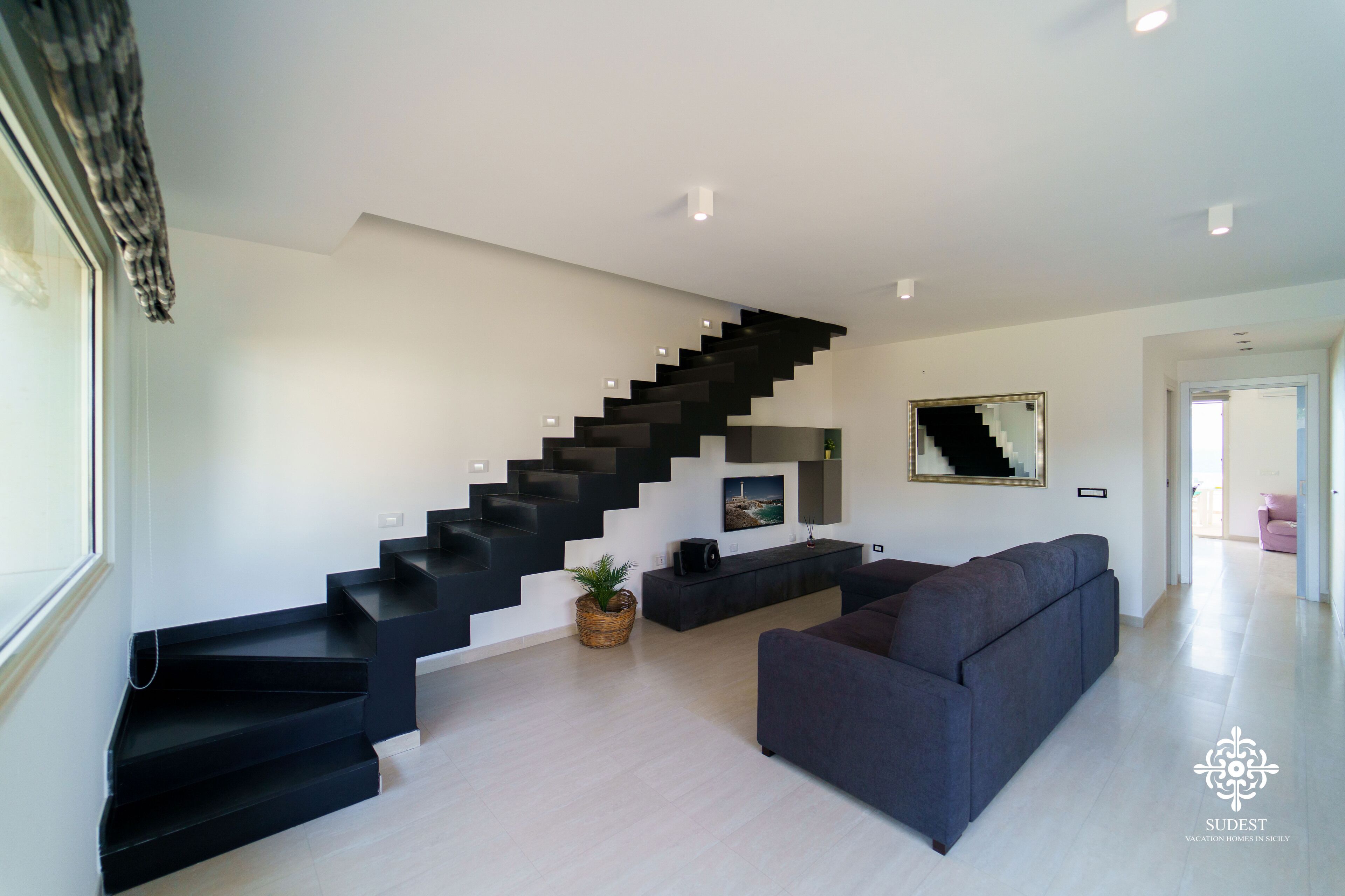 A modern living room with a black staircase, a grey sofa, and a flat-screen TV on a black console.