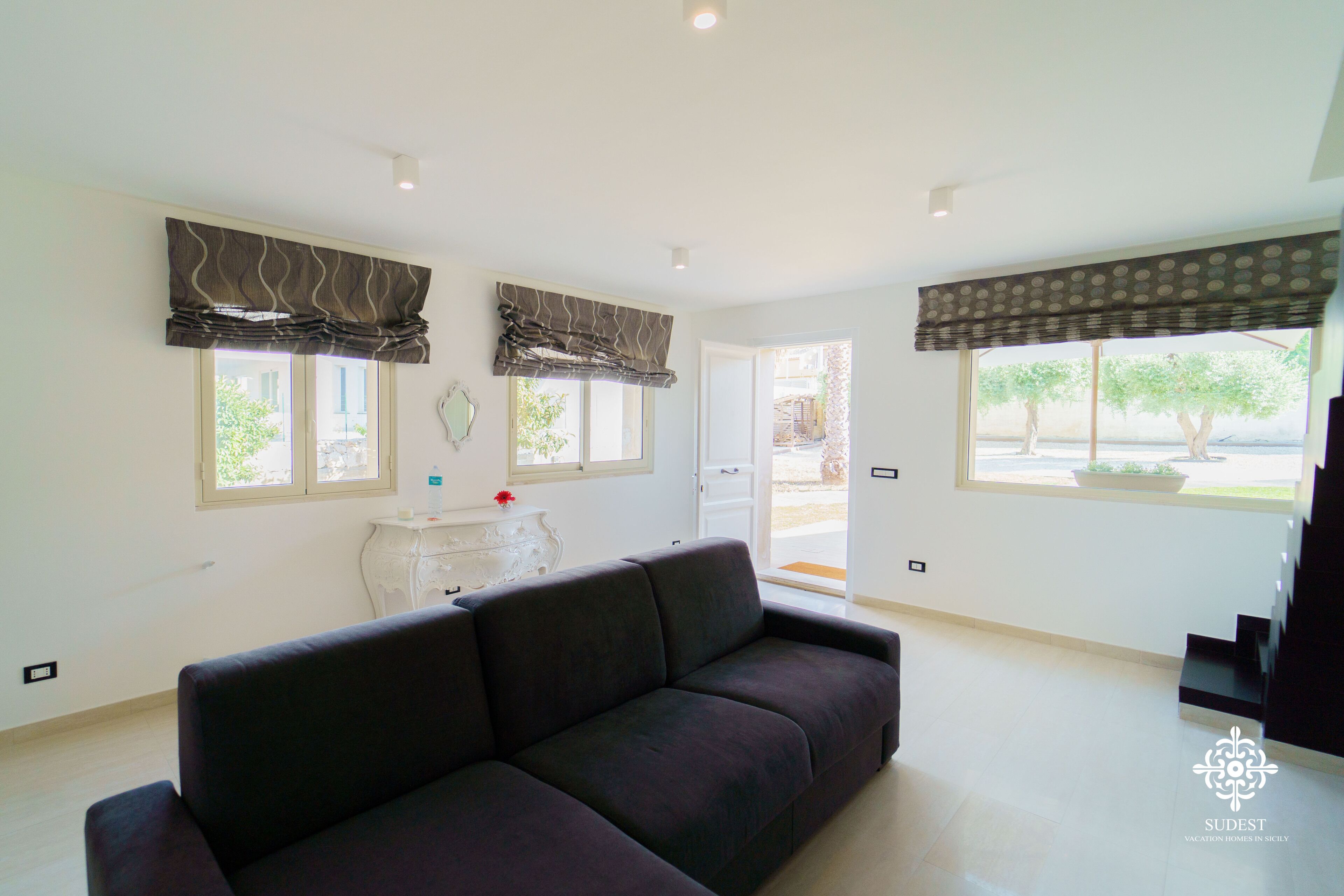 A modern living room with a dark sofa, large windows with curtains, a white side table, and a staircase leading to an upper level.