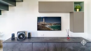 A modern living room with a large flat-screen TV mounted on the wall, a dark wooden cabinet below, and a small green shelf with a plant. - Villa Vittoria  - Dimora fronte mare (Augusta)