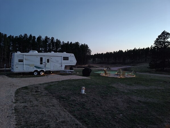 A white RV parked in a grassy area with a picnic table and chairs, surrounded by trees.