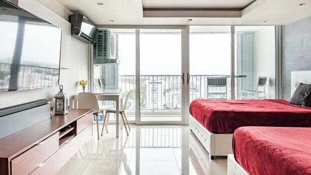 A modern hotel room with a large bed, a TV, a desk, and a balcony with a city view.