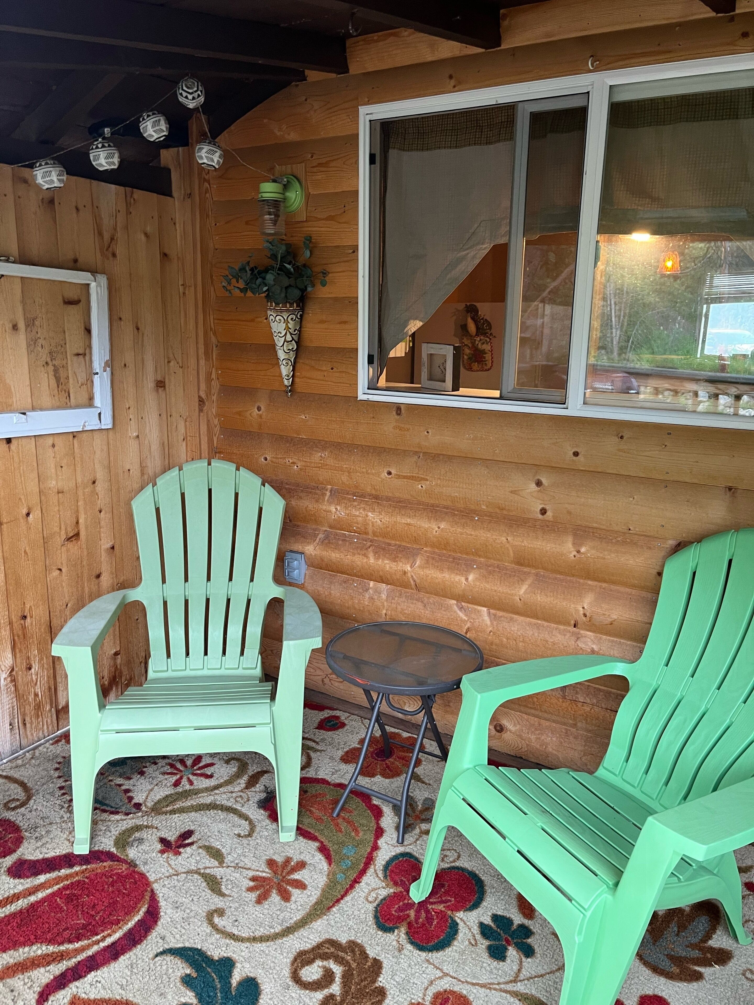 A wooden cabin interior with two green Adirondack chairs, a small round table, and a window with a view of the outside.