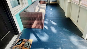 Eine überdachte Veranda mit blauem Boden, weißem Zierrat und Blick auf die Straße. Zwei Korbstühle und eine Couch mit Kissen sind vorhanden.
