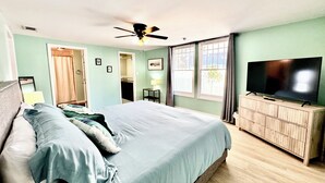 Ein Schlafzimmer mit einem Bett, einem Deckenventilator, einem Fernseher auf einer Holzkommode und einem Fenster mit Vorhängen.