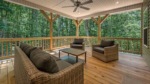 A covered outdoor deck with wooden flooring, wicker furniture, and a ceiling fan. - Fawn'd Memories - Hot Tub | Fire Pit | Gas Grill (Blairsville)