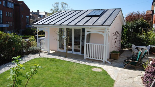 Et orangeri med glasfacade, en hvid veranda og en have med græsplæne og plantekrukker.