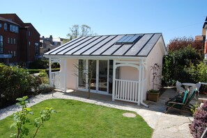 Une véranda avec une structure en verre, une terrasse blanche et un jardin agrémenté d’une pelouse et de plantes en pot.