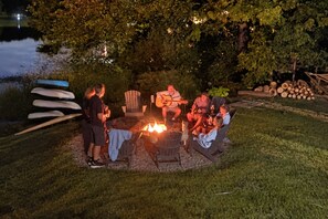 An outdoor gathering around a fire pit with people seated and standing, surrounded by trees and a body of water.