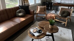 Una sala de estar moderna con un sofá de cuero marrón, dos sillas de madera, una mesa de centro con una planta y una cocina al fondo.
