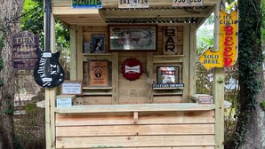 Un bar exterior de madera con techo de láminas, con letreros que dicen 'The Ben Backyard Bar' y 'Beer Bullshit'. Hay tres taburetes de metal adelante.