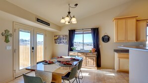 Un comedor con una mesa, sillas y un candelabro. Hay un ventanal con cortinas, una zona de cocina con armarios, y una puerta que da al exterior.