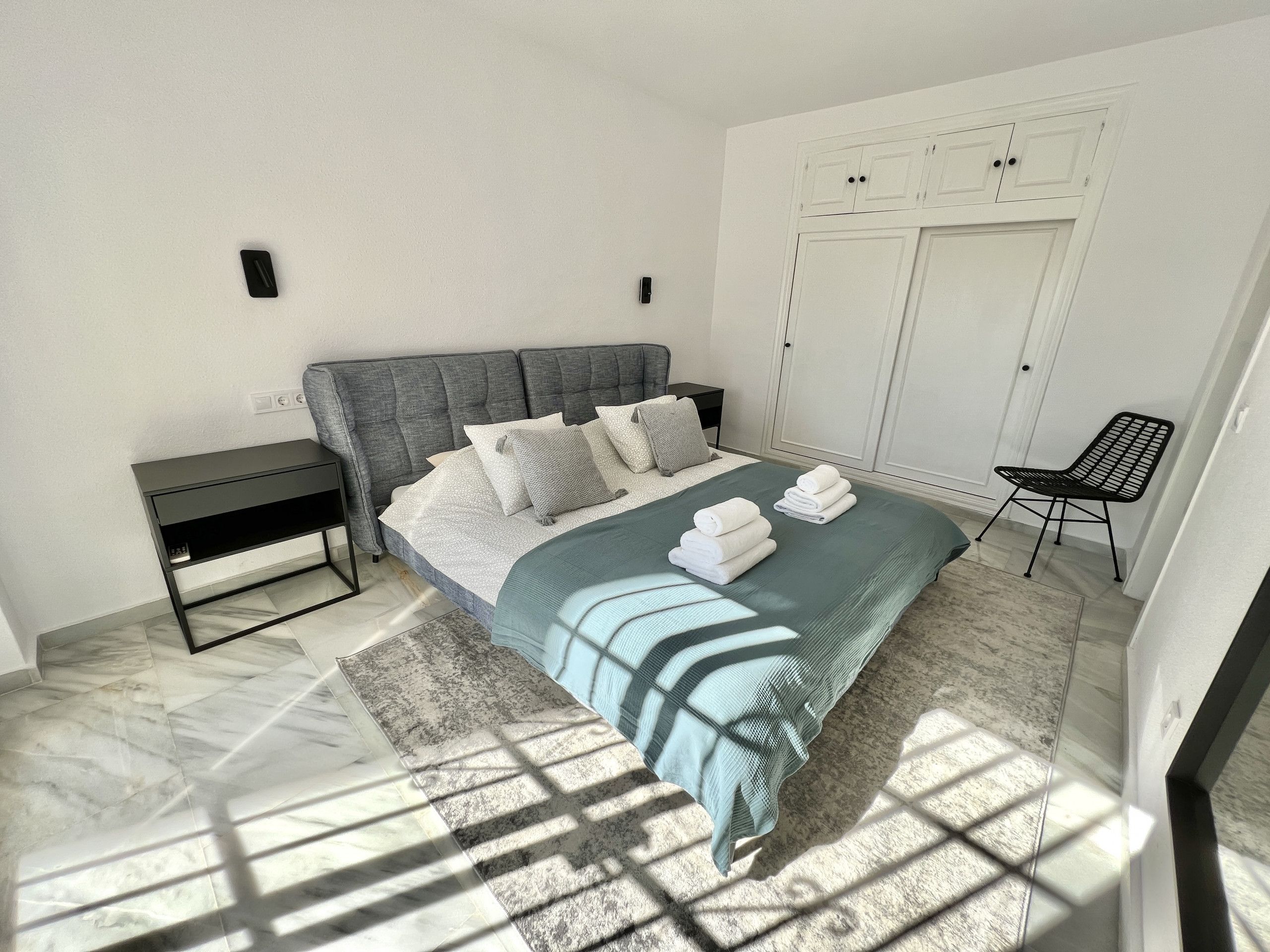 A modern bedroom with a grey upholstered headboard, a bed with grey and white bedding, a black metal chair, and a white wardrobe.