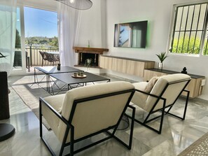 A modern living room with a fireplace, a dining area, and a large window with a view of greenery.