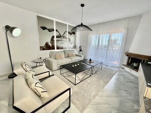 A modern living room with a sofa, coffee table, and floor lamp. There is a fireplace and a large window with curtains.