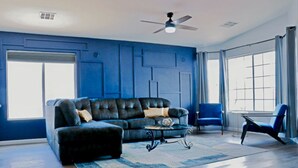 A living room with a dark leather sofa, a coffee table, and a blue armchair. There is a ceiling fan and a window with curtains.