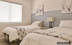 Une deuxième chambre avec deux lits, une fenêtre avec des stores, une table de chevet avec une lampe et des œuvres d’art abstraites accrochées au mur.