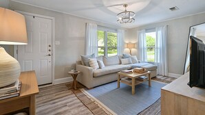 A living room with a sofa, coffee table, and TV. There is a door and a window with curtains. - Enjoy our cozy cottage just minutes from A&M Campus (Bryan)