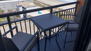 A balcony with two chairs and a table, overlooking a beach with a parking lot and buildings in the background.