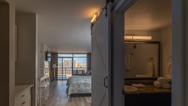 A hotel room with a bed, a bathroom with a mirror and towels, and a view of a cityscape through large windows.