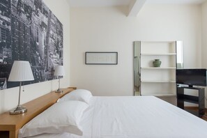 A hotel room with a bed, two bedside lamps, a framed picture, a television on a stand, and a large cityscape painting on the wall.