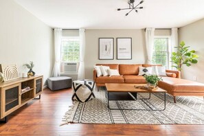 A modern living room with a brown sofa, a coffee table, and a patterned rug. There are two framed pictures on the wall and a plant on the right. - Spacious Lake Community House near Hudson (Athens)