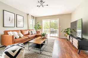 A modern living room with a brown leather sofa, a wooden coffee table, and a black TV stand. There are two framed pictures on the wall and a large window with a view of greenery outside. - Spacious Lake Community House near Hudson (Athens)