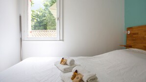 A neatly made bed with white linens and towels, a wooden headboard, and a window with a view of trees and a tiled roof. - Appartement Pompidou - Welkeys (La Seyne-sur-Mer)